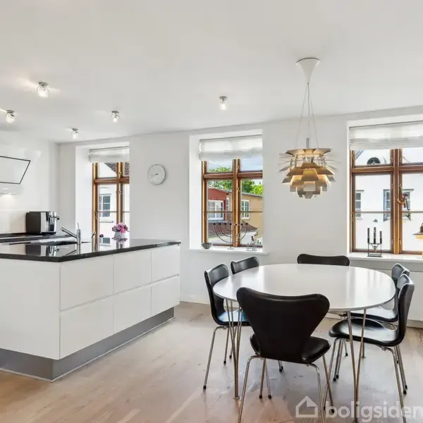 Et moderne køkken er indrettet med en stor køkkenø, en rund spisebord med sorte stole og en designerlampe. Rummet er lyst med trægulve og tre store vinduer.