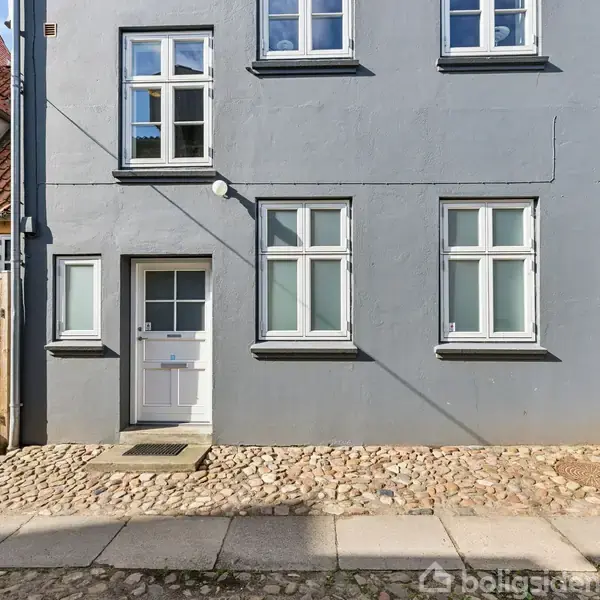 Grå bygning med hvid dør og vinduer, står stille på brostensbelagt fortov; omgivet af nabohuse med rødt tegltag og træhegn.