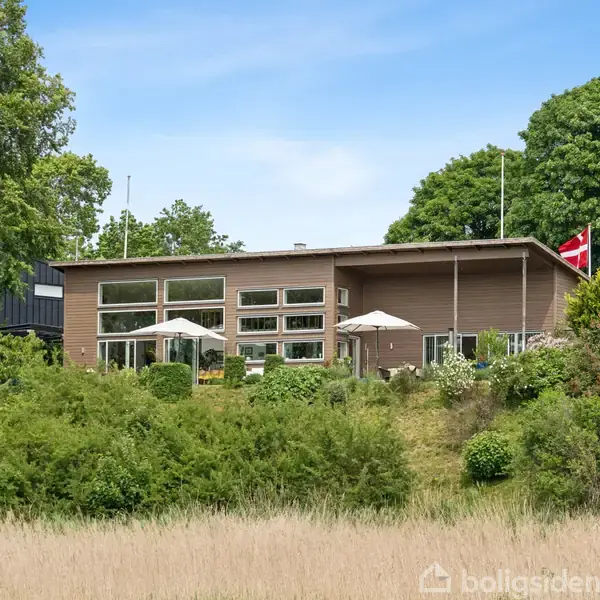 Hus med moderne arkitektur, har store vinduer og dansk flag. Udenfor står parasoller på terrassen. Omgivet af frodig vegetation og grønne træer.