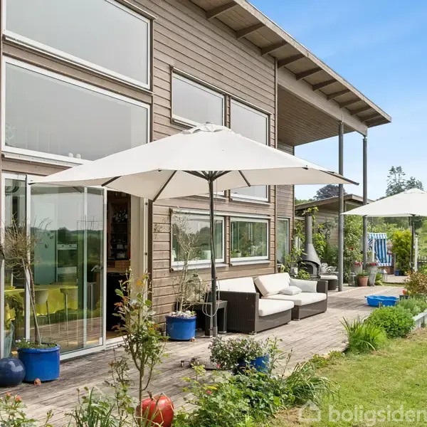 Moderne træhus med store vinduer, sofa og parasoller på terrasse omgivet af blomsterkrukker. Græsplæne og grønne planter udgør en frodig have.