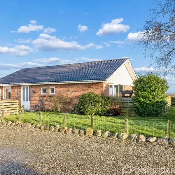 En murstenshus med mørkt tag stående på en velplejet græsplæne, omgivet af buske og et trægærde. Ved siden af ligger en lille træbygning. Dagen er klar og solrig.