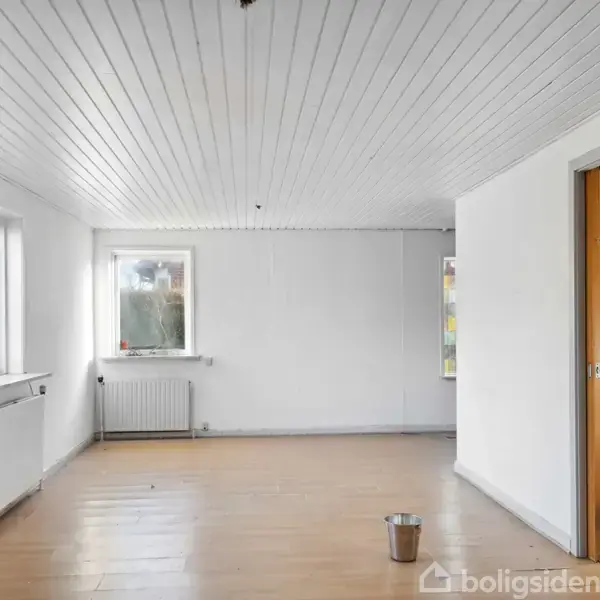 Et tomt rum med hvide vægge og loft, trægulv, og store vinduer til venstre. En metalspand står på gulvet. Rummet har en opvarmningsradiator og en åben dør.