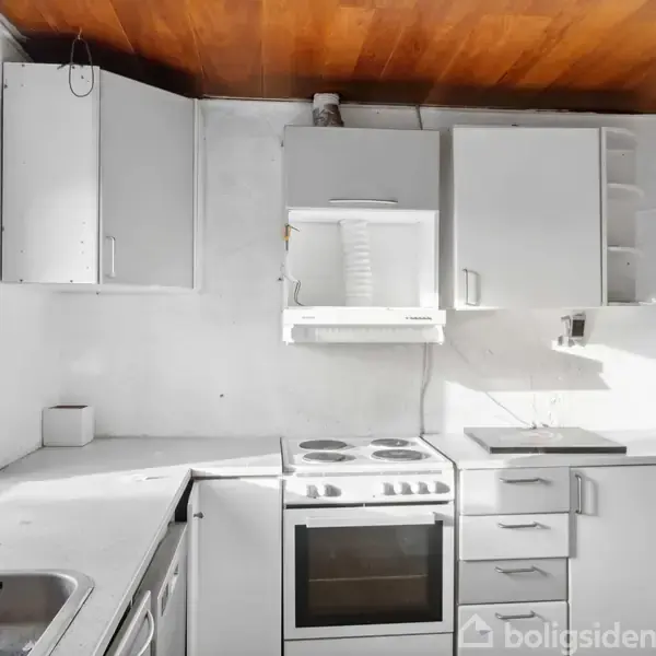 Et funktionelt køkken med hvidt interiør, herunder komfur, emhætte og flere skabe. Sollys skinner ind gennem vinduet til venstre, og loftet er i en varm træfarve.