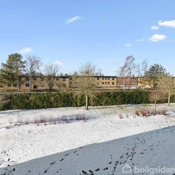 Snebeklædt have med fodspor ses i forgrunden, omgivet af nogle bare træer. I baggrunden findes en række bygninger og buske, omgivet af en blå himmel med få skyer.