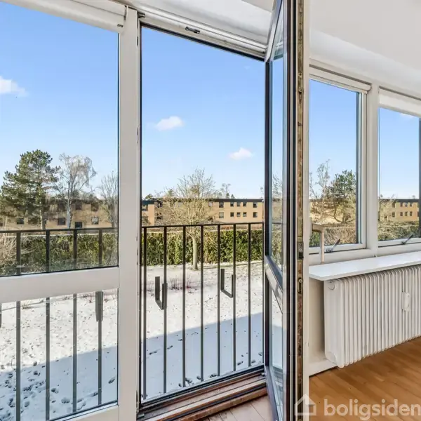 En glasdør står åben mod en balkon med gelænder, hvorfra der er udsigt til træer og bygninger under en blå himmel. Indendørs ses et radiatorskjuler ved trægulv.