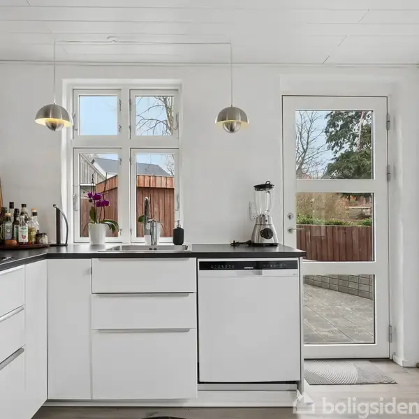 Et moderne køkken med hvide skabe, hvor der står en opvaskemaskine. En blender står på bordet. Gennem vinduet og døren ses en flisebelagt terrasse og træhegn udenfor.