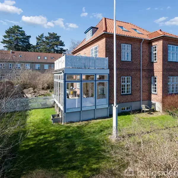 Rødt murstenshus står på en grøn græsplæne, med store glaspartier på tilbygning. Omkring huset er en have med buske og træer, under en blå himmel med spredte skyer.