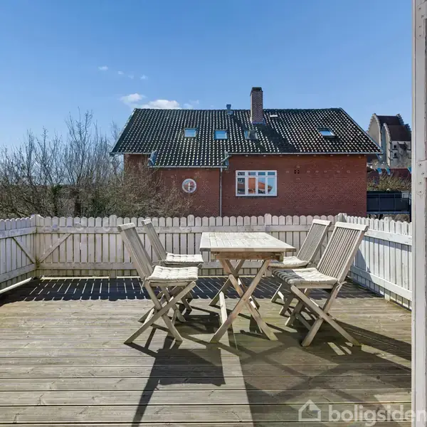 Terrassemøbler står på en træterrasse, omgivet af et hvidt stakit. I baggrunden ses et træbeklædt hus og en blå himmel.