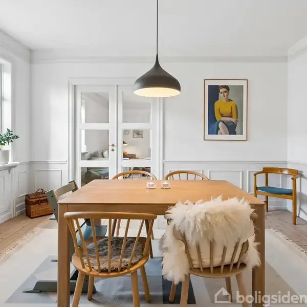 Spisebord med stole står centralt under en sort pendellampe i et lyst rum med trægulve. Væggene er hvide med et maleri, og vinduer giver lys.