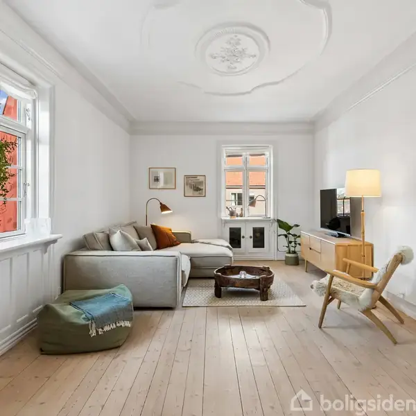 Stue med sofa og bord i handling, omgivet af hvide vægge og trægulv. En grøn puf, bogreol, og planter skaber en indbydende atmosfære. Lampe og vinduer tilfører lys.