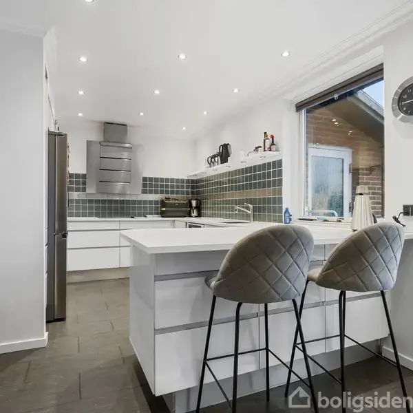 Et moderne køkken med hvide skabe og mørke fliser viser en ø med to polstrede barstole. En stor emhætte er over komfuret. Et ur hænger på væggen ved siden af en glasdør.