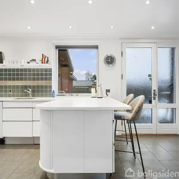 Et moderne køkken med en stor hvid ø, barstole og flisegulv. Hylder med køkkenredskaber hænger over en flisebeklædt væg. En glasdør og vindue bringer lys ind i rummet.