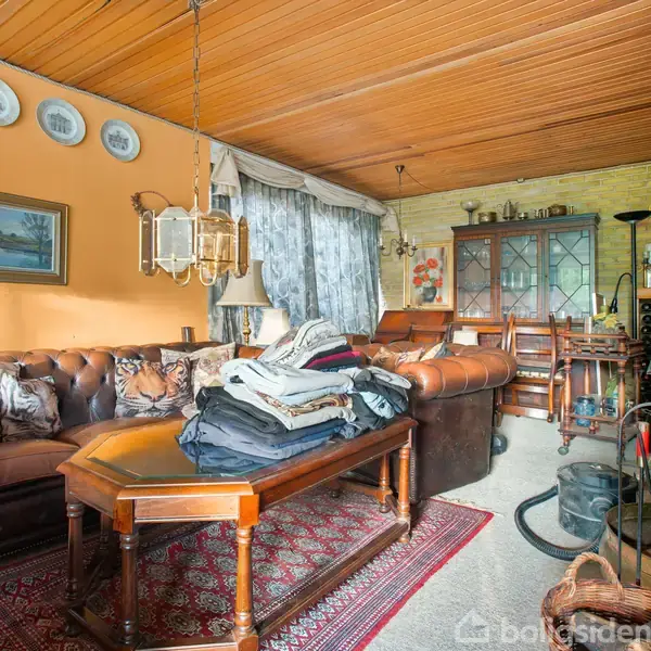 Brunt læderhjørnesofa med puder omgivet af træmøbler i en stue. Mørkerødt tæppe på gulvet. Høj, fyldt reol i baggrunden. Tøj stablet på sofabordet. Lysarmatur hænger fra loftet.