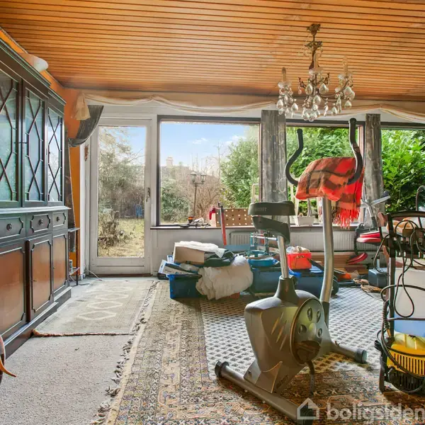 En motionscykel står i en stue fyldt med møbler og tæpper. Et skab langs væggen til venstre. Store vinduer viser en have udenfor. En lysekrone hænger i loftet.