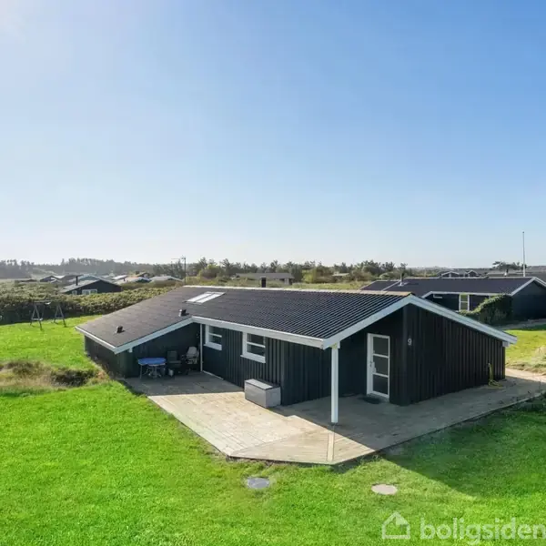 Et sort, en-etagers hus med terrasse ligger på en stor græsplæne. Huset er i et beboelsesområde med flere lignende bygninger omkring. Solen skinner fra en klar himmel.