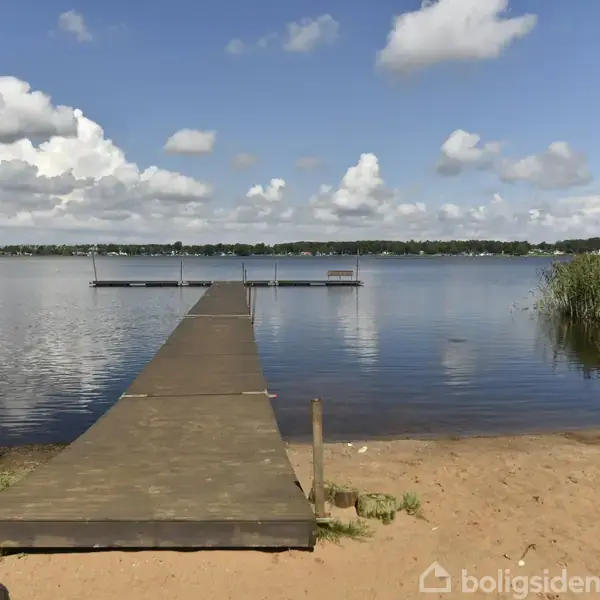 En træbrygge strækker sig over en rolig sø mod en fjern kystlinje. Skyer pryder en blå himmel, og bredderne er omgivet af siv og sand.