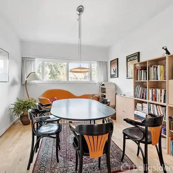 Et spisebord med seks sorte stole står i et lyst rum. En orange sofa og reol med bøger er i baggrunden. En lampe hænger over bordet, og der er en plante ved vinduet.