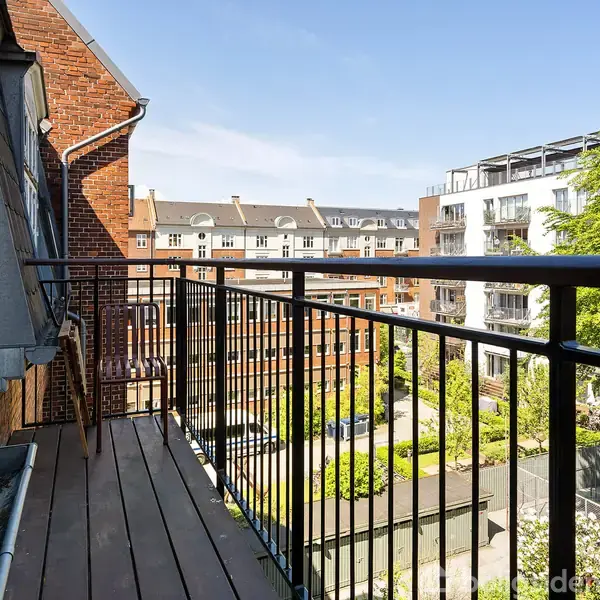 altan med trædækning og metalrækværk, som tilbyder udsigt over en gårdhave omgivet af murstensbygninger. Grønne træer og klare blå himmel i baggrunden.
