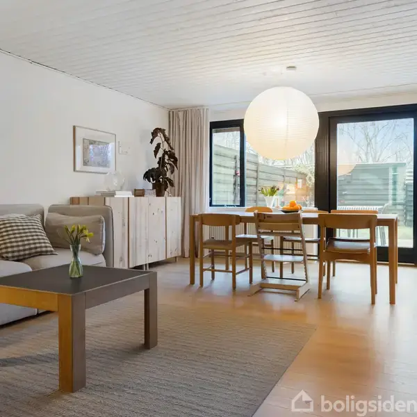 En stue har en grå sofa ved siden af et sofabord og spisebord med seks stole. Store vinduer i baggrunden giver udsigt til en have. En lampeskærm hænger over bordet.