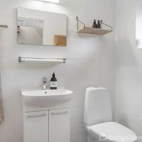 Håndvask med spejl hænger på en hvid væg. Toiletbesætning til højre under et lille vindue. Håndklæde hænger ved siden af døren i et lyst, minimalistisk badeværelse.