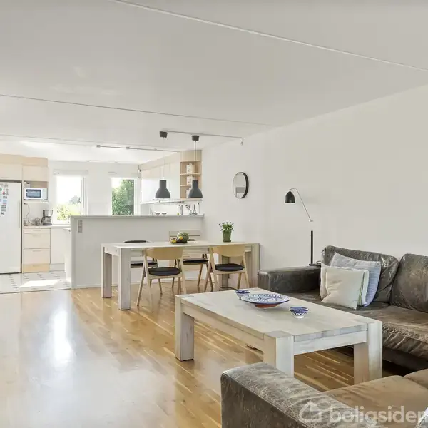 En stue med to brune sofaer og et sofabord. Ved siden af er et spisebord med fire stole. I baggrunden ses et åbent køkken med hvidt interiør.