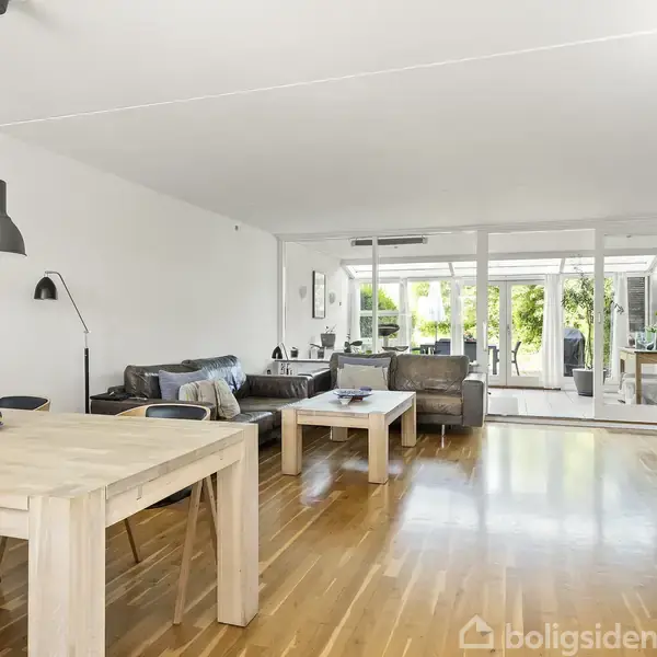 Et lyst og moderne stueområde med træmøbler. Et spisebord med frugter står i forgrunden, sofagruppe bagerst. Store vinduer viser en have udenfor.
