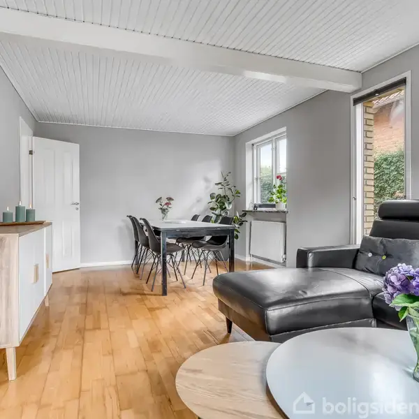 En sort lædersofa står i en stue med trægulv og hvide vægge; sofabord med en vase med lilla blomster; spisebord i baggrunden; vinduer med planter giver naturligt lys.