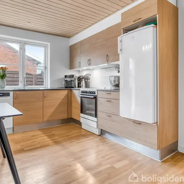 Køkken med træskabe og hvidevarer, herunder komfur og køleskab, står stille i et lyst rum med trægulv og en lampeovn på bordet. Et vindue giver dagslys ind.