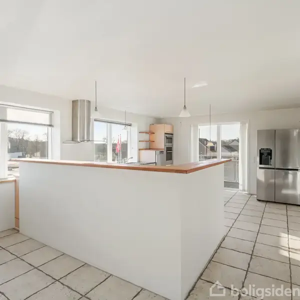 Køkken med hvidt interiør og køleskab, rummelige vinduer giver naturligt lys, set i en moderne bolig med flisegulv. På vinduet ses skiltet "Til salg".
