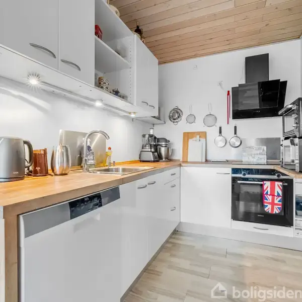 Et moderne køkken med træbordplade og hvide skabe, indeholder en vask, elkedel og diverse køkkenudstyr. Ovn med et viskestykke hængende. Lyse træloft og beige gulv.