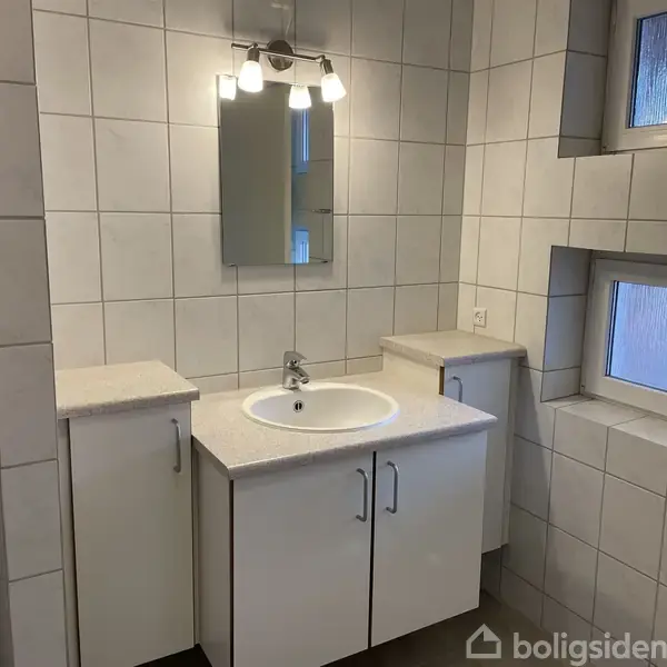 Håndvask og spejl oplyst af lampe, omgivet af hvide fliser. To vinduer på væggen til højre. Modernistisk badeværelsesindretning med enkle, hvide underskabe.