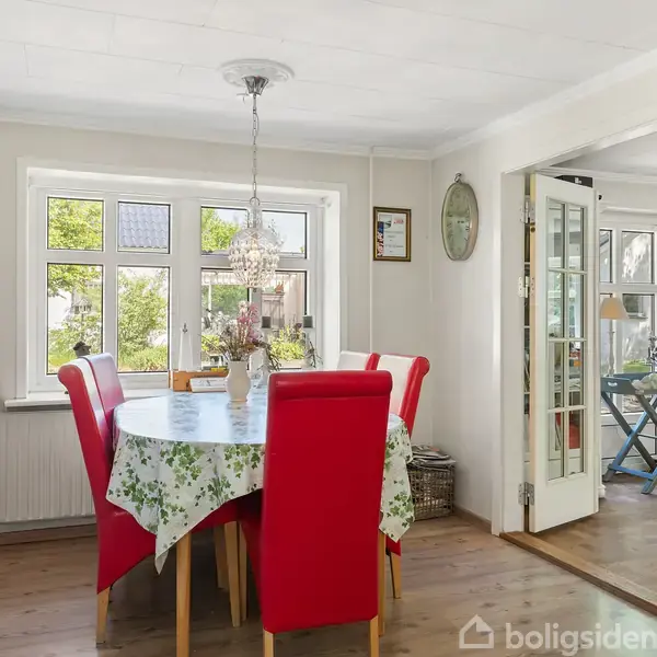 Rundt spisebord med blomstret dug omgivet af røde stole. Bordet står i et lyst og hyggeligt spisestueområde med vinduer og franske døre, der åbner til stue med lænestol.
