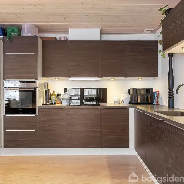 Køkken med mørke træskabe, hvidevarer som ovn og mikrobølgeovn. Køleskab med magneter til venstre. Trægulv og -loft. Indbygningsbelysning under skabe. Vandhane ved vask til højre.