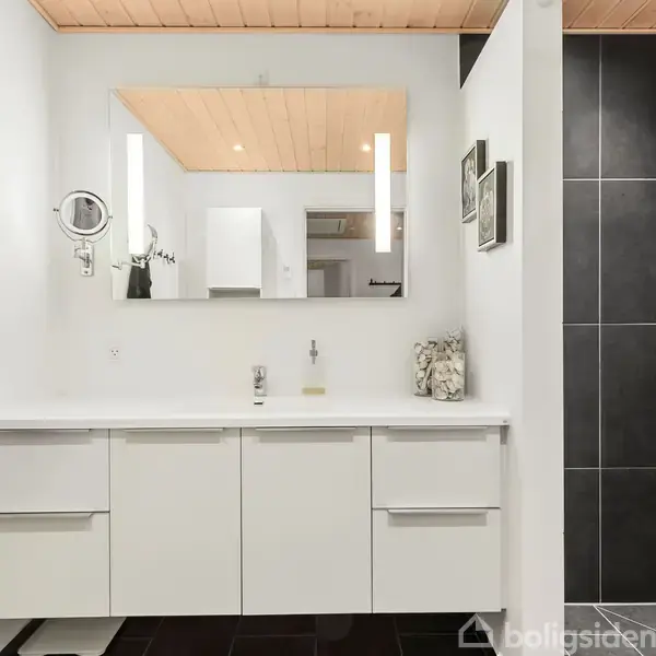 Et hvidt badeværelsesmøbel står under et stort spejl. En grå håndklæde hænger ved siden af. Til højre er der en bruser med mørke fliser. Loftet er træ.