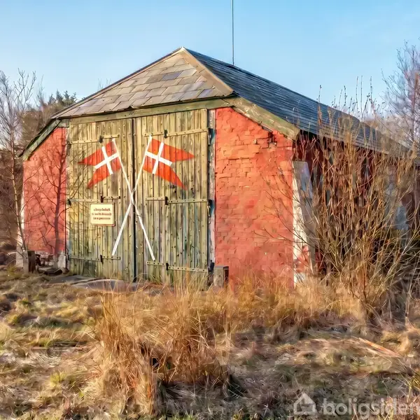 En rød bygning med grønne porte, prydet med to danske flag. Bygningen er omgivet af vildtvoksende græs og træer. To skilte på portene viser teksten, men teksten er utydelig.