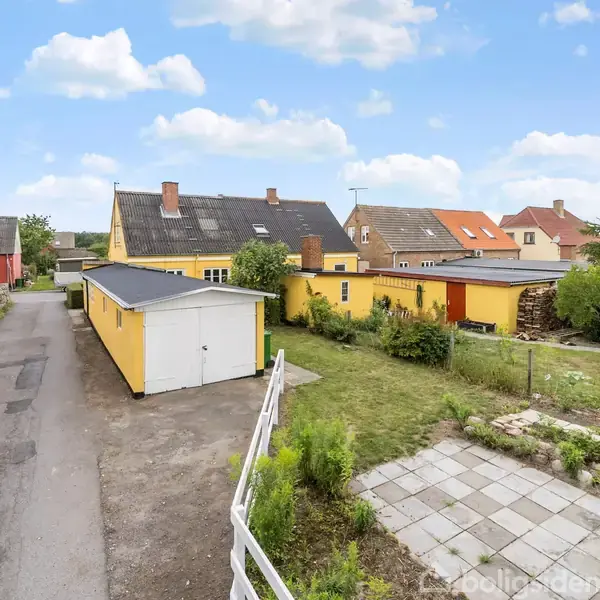 Et gult hus står ved siden af en garage og have, placeret i et roligt kvarter med flere farverige huse og et stenbelagt fortov langs vejen.