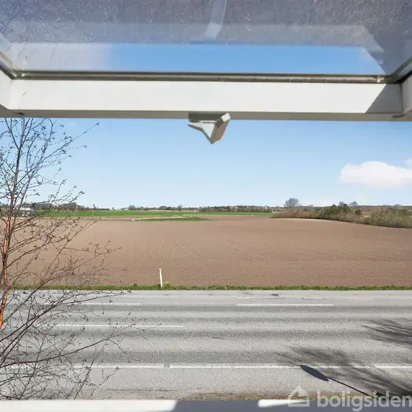Et åbent vindue giver udsigt til en vej og en brun mark under en klar blå himmel. Grønne marker og træer vises i baggrunden, der harmonerer med den fredfyldte landskabsudsigt.