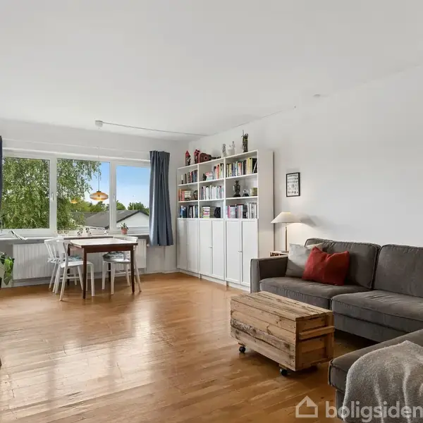 Gråt hjørnesofa stående i en lys stue med træbord, reol fyldt med bøger og et stort vindue, der giver udsigt til grønt område. Plankegulve og simple dekorationer skaber en hyggelig atmosfære.