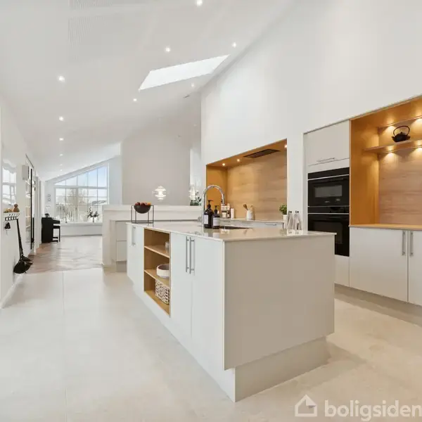 Et moderne køkken med lysegrå skabe og en central ø. Loftbelysning lyser rummet op, med åbne hylder og indbyggede ovne. I baggrunden ses en stue med store vinduer.