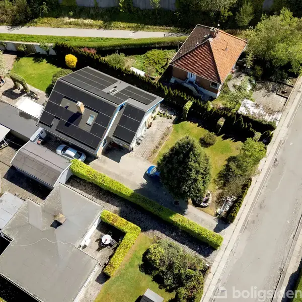 Et parcelhus med sort tag set fra oven. Huset er omgivet af en grøn have med træer og buske. Det ligger i et boligområde med en vej ved siden af.