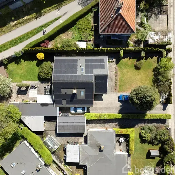 Hus med solpaneler på taget, set fra oven, omgivet af haver og indkørsel med en blå bil. Beliggende i et boligkvarter med stier, veje og flere bygninger i nærheden.