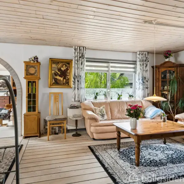 En stue med lyse trægulve har en beige hjørnesofa og et sofabord med en blomstervase. Rummet indeholder også et vitrineskab, vægkunst og åbner sig mod en spisestue gennem en bue.