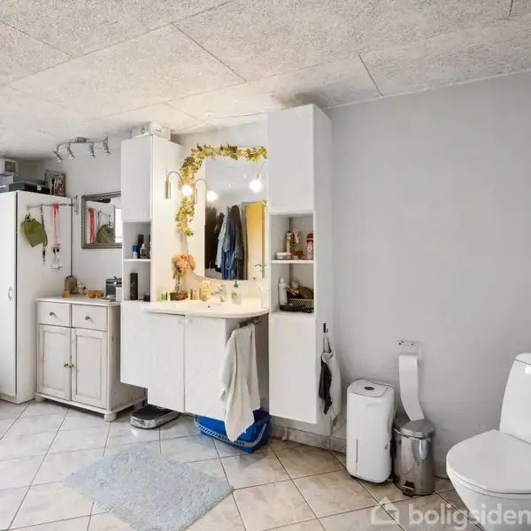Badeværelse med hvidt skabsarrangement; spejl omkranset af lys og grønne ranker. Toilet til højre, skraldespand og opbevaring. Lysegråt gulv med firkantede fliser.
