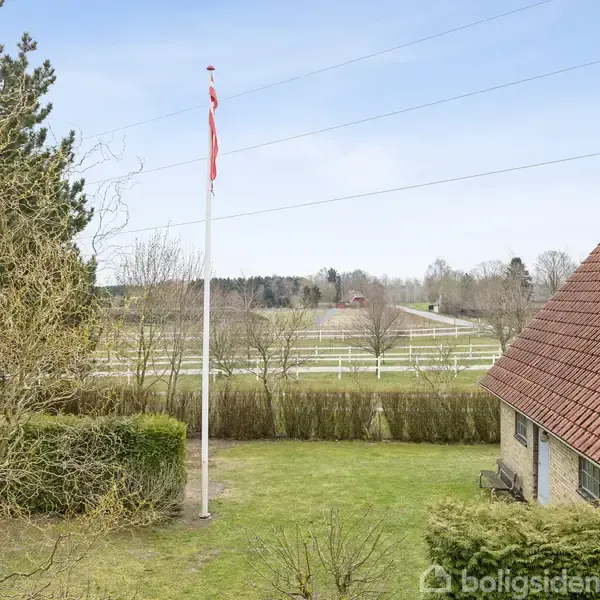 Flagstang med dansk flag står i ro på en grøn græsplæne ved siden af et hus med rødt tag, omgivet af hække og åbne marker i baggrunden.