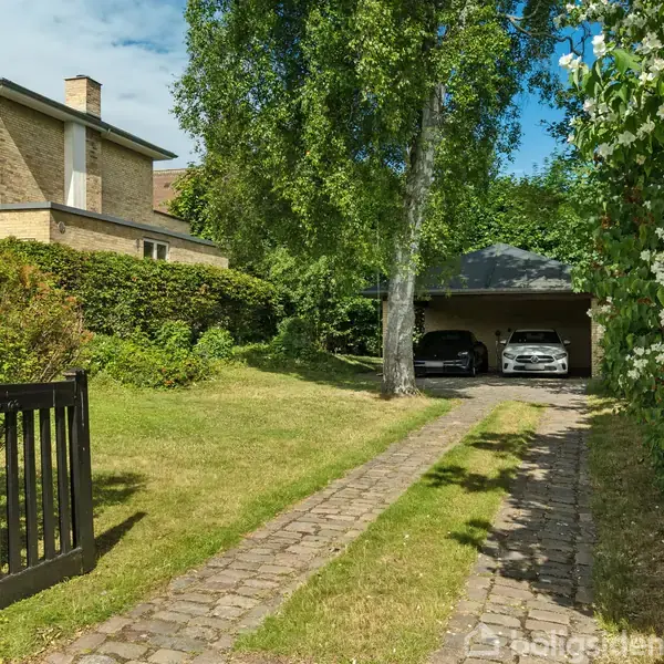 Et tofamiliehus i baggrunden, en græsset indkørsel med brosten i forgrunden, to biler parkeret i en overdækket garage til højre, omgivet af grønne buske og træer.