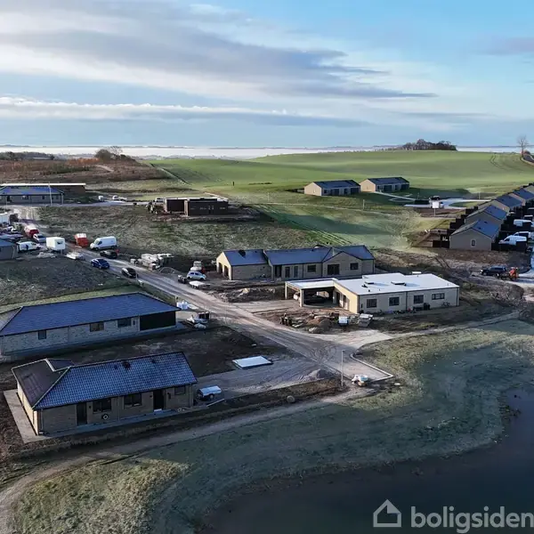 Flere nybyggede huse står langs en vej i et åbent, landligt område nær en lille sø. Andre bygninger er synlige i baggrunden, omgivet af marker og åbne vidder.