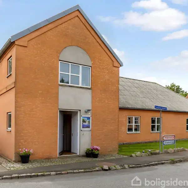 Bygning i rød mursten med en åben dør står langs en stille vej. Der er græsplæne og buske. Et skilt foran viser vejnavnet “Stationsvej”.