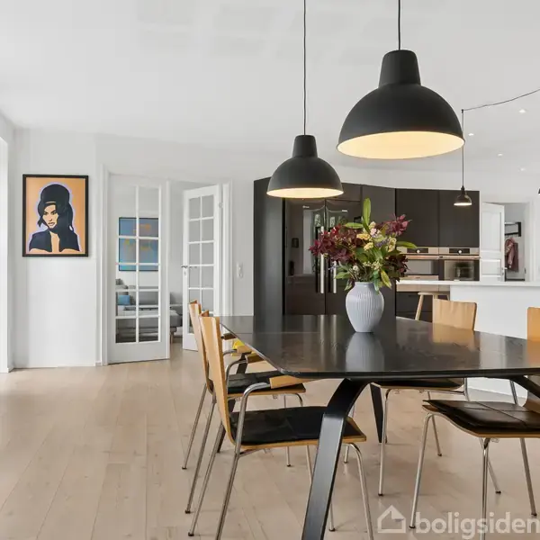 Et sort spisebord med træstole står i et lyst rum, dekoreret med en vase blomster. Loftslamper hænger over bordet. I baggrunden ses et åbent køkken og en dør med en plakat.