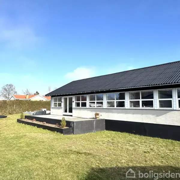 En bungalow med sort tag og hvide vinduer står på en stor græsplæne omgivet af hække og nogle få træer under en klar blå himmel.