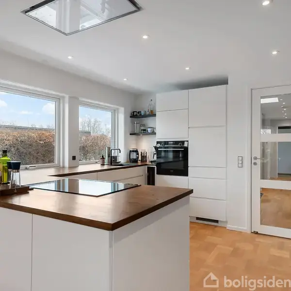 Et moderne køkken med hvide skabe og træbordplade, indeholder kogeplade, køkkenmaskiner og vinduer, der giver udsigt til en hæk. Et parketgulv og en dør fører til et andet rum.